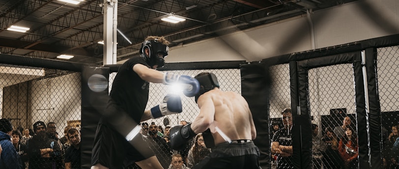 Two fighters engage in a mixed martial arts match within a caged ring. They are wearing protective gear, including gloves and shin guards, and are surrounded by spectators watching intently. Harsh lighting from above casts shadows on the fighters and the mat.